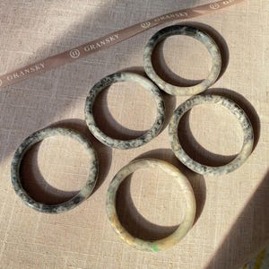 May include: Five jade bangles with a mottled black and white pattern. The bangles are stacked on a beige fabric surface.