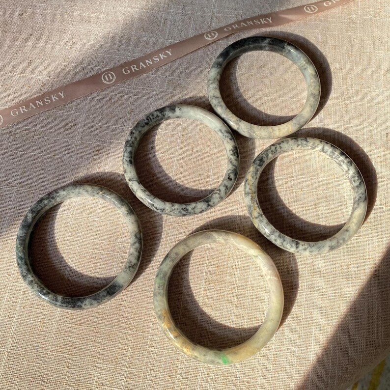 May include: Five jade bangles with a mottled black and white pattern. The bangles are stacked on a beige fabric surface.