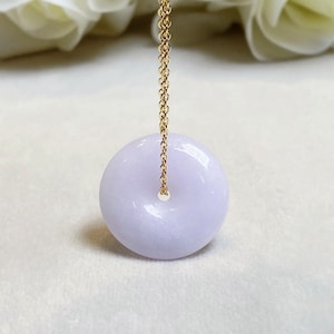 May include: A light purple jade donut pendant with a gold chain. The pendant is round with a hole in the center. The gold chain is delicate and attached to the top of the pendant. The background is a soft white.
