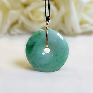 Könnte beinhalten: Ein Jade-Anhänger in kreisförmiger, donutartiger Form. Der Anhänger hat helle und dunkle Grüntöne, einen goldfarbenen Metallakzent und eine schwarze Kordel. Der Anhänger hängt vor einem weichen, verschwommenen Hintergrund.