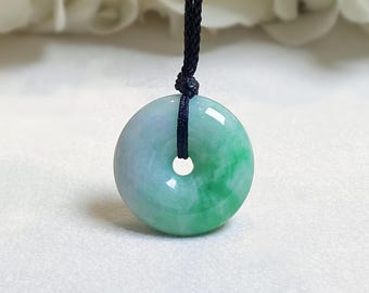 May include: A circular jade pendant with a central hole, hanging from a black cord. The pendant displays a mix of light green and pale blue colours, with a polished surface. The backdrop is a soft, white fabric.