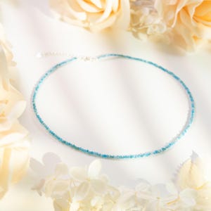 May include: A delicate blue gemstone necklace with a silver clasp. The necklace is made of small, faceted beads and is displayed on a white background with flowers.