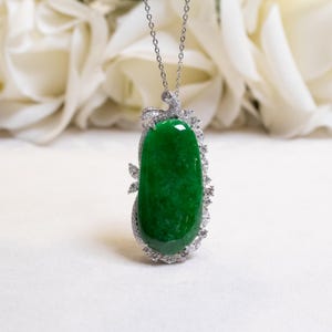 Può includere: Una collana con pendente di giada con una vibrante pietra di giada verde. La giada è incastonata in una montatura color argento ornata di piccole gemme trasparenti. La collana è sospesa da una sottile catena d'argento, che presenta un elegante gioiello.