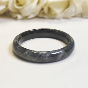 May include: A dark gray jade bangle bracelet. The polished bracelet has a smooth, rounded surface with subtle variations in color and texture. The jade is a mix of dark gray and lighter gray tones, with a glossy finish.
