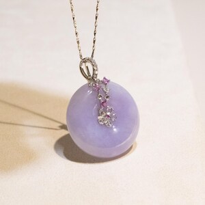 May include: A lavender-colored pendant necklace with a silver chain. The pendant is a smooth, circular stone with a floral design of pink and clear gemstones. The necklace is displayed on a white surface.