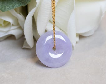 May include: A lavender jade donut pendant with a gold chain. The polished stone has a circular shape with a hole in the centre. The pendant hangs in front of a white flower.