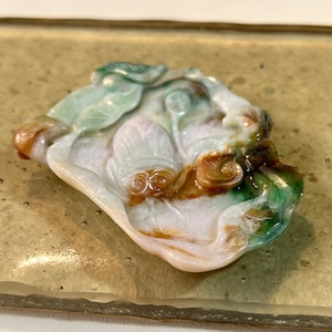 May include: A jade pendant carved with a detailed design of two frogs sitting on a lotus leaf. The pendant is a mix of green, white, and brown jade.