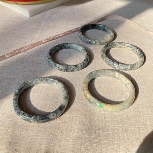 May include: Five jade bangles in various shades of green and black with white marbling. The bangles are arranged on a light beige fabric surface.