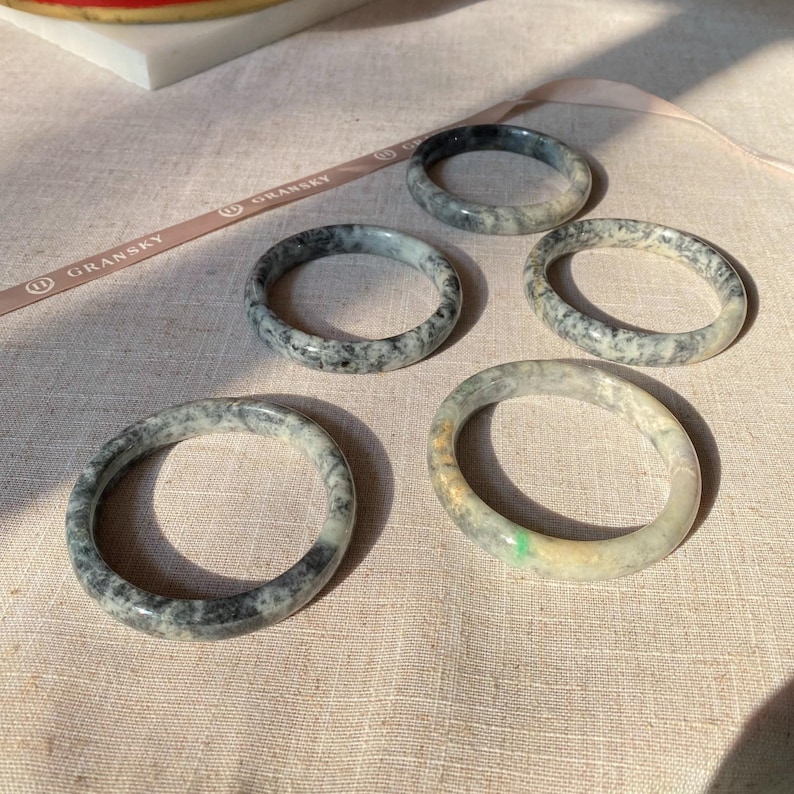 May include: Five jade bangles in various shades of green and black with white marbling. The bangles are arranged on a light beige fabric surface.