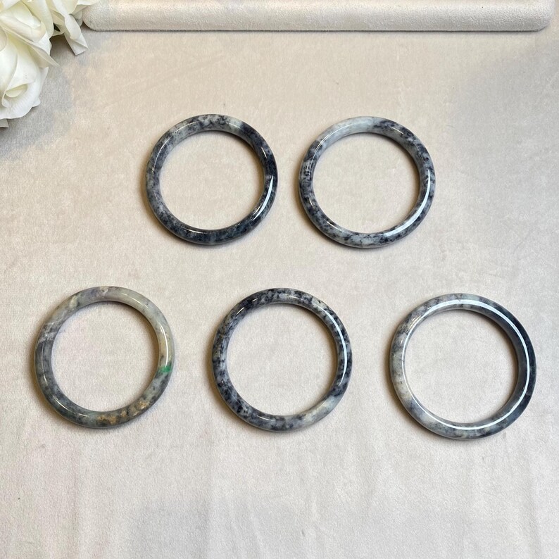 May include: Five jade bangles with a mottled black and white pattern. The bangles are arranged in a semi-circle on a white surface.