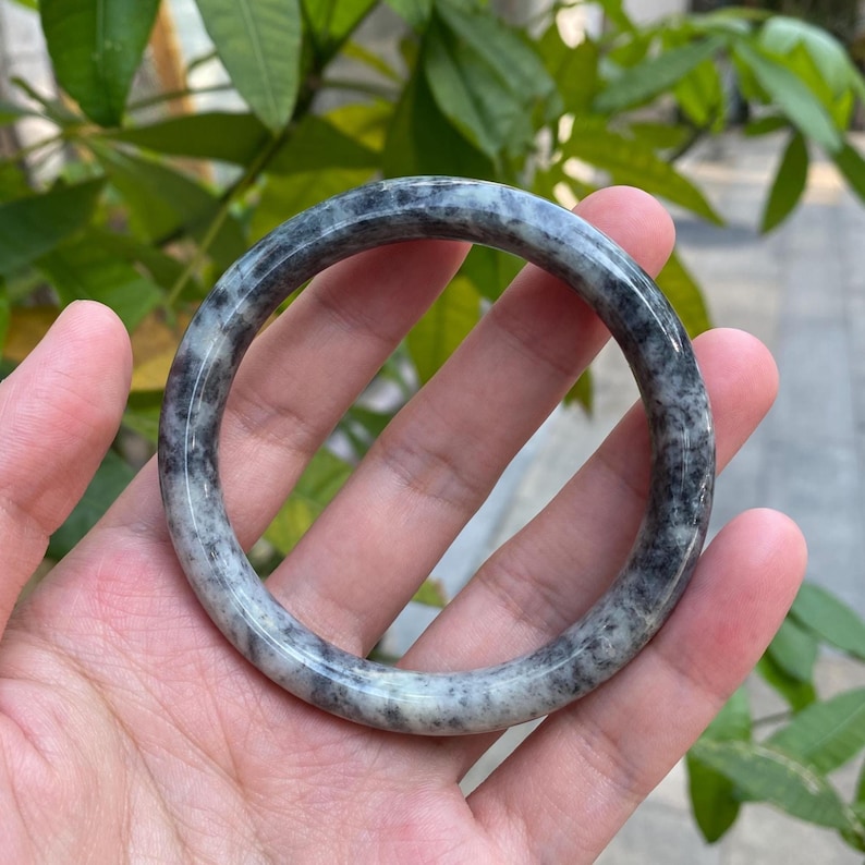 May include: A gray and white jade bangle bracelet with black speckles. The bangle is smooth and polished.