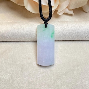 May include: A rectangular jade pendant with a gradient of green, white, and lavender hues, suspended from a black cord. The jade has a polished surface and a smooth texture. The pendant is a piece of jewelry.