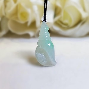May include: A pale green, translucent, carved stone pendant shaped like a spiral seashell. The pendant features a smooth, polished surface with subtle color variations and is suspended from a thin, black cord that is tied at the top of the shell.