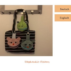 Könnte beinhalten: Eine schwarz-weiß gestreifte Tragetasche mit drei daran befestigten Reißverschlusstaschen in Form einer Katze, eines Bären und eines Frosches. Die Tasche hängt an einer Wand mit einem Metallgestell.