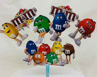 May include: A cake topper featuring nine colorful M&M's characters. The characters are all smiling and have different expressions. The topper is made of cardboard and has a blue plastic stick for inserting into a cake.