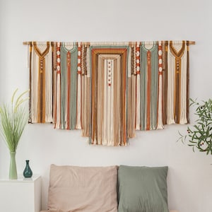 May include: A large, colorful macrame wall hanging with a bohemian style. The wall hanging is made with natural fibers and features a variety of textures and patterns. It is hanging above a bed with beige and green bedding.