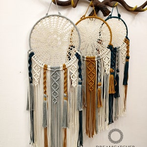 May include: Three macrame dreamcatchers with white, gray, teal, and mustard yellow tassels hanging from a wooden antler rack. The dreamcatchers have a white crocheted center and are decorated with different macrame patterns. The text "Dreamcatcher Store" is visible at the bottom of the image.
