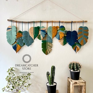 May include: A wall hanging made of macrame with a tropical leaf design in shades of green, blue, and yellow. The hanging is made of yarn and is attached to a wooden branch. The text "DREAMCATCHER STORE" and "HANDMADE BY NHAM" is visible below the hanging.