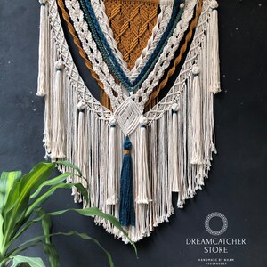 May include: A large macrame wall hanging with a triangular design. The macrame is made with natural colored cord and features accents of teal, mustard yellow, and white. The wall hanging is attached to a wooden dowel.