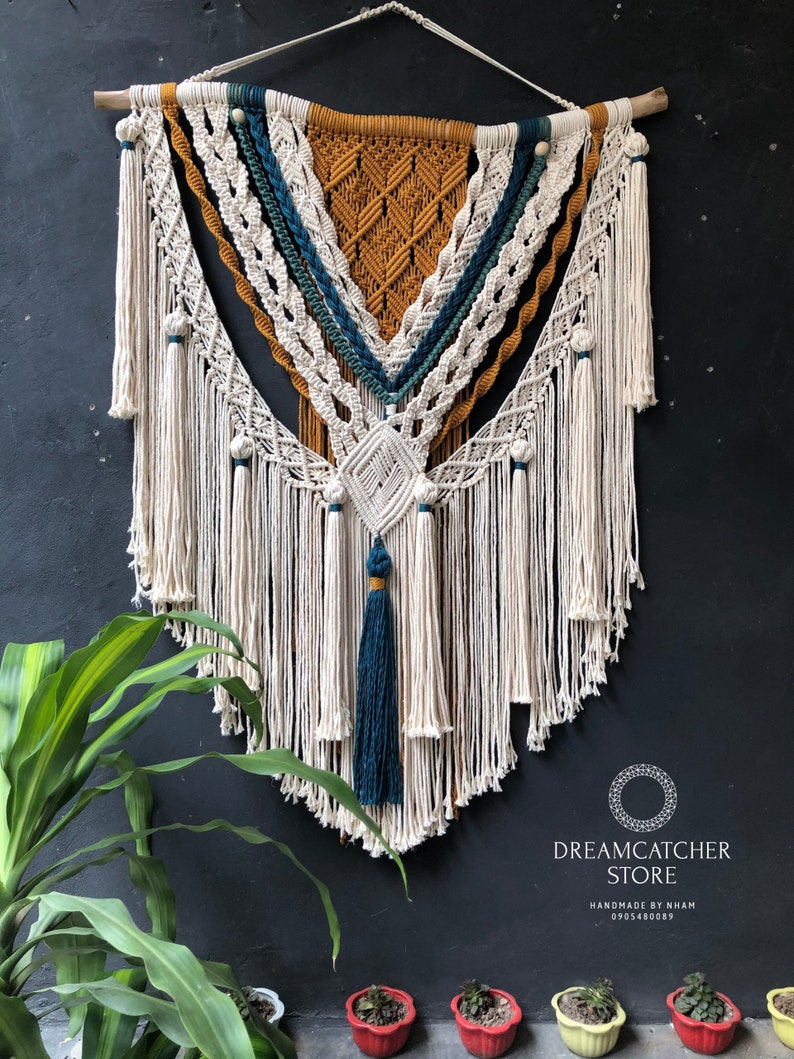 May include: A large macrame wall hanging with a triangular design. The macrame is made with natural colored cord and features accents of teal, mustard yellow, and white. The wall hanging is attached to a wooden dowel.