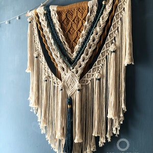 May include: A large, beige macrame wall hanging with a geometric pattern in shades of brown, green, and blue. The hanging is made with thick cotton cord and has a wooden dowel at the top. The hanging is decorated with small white beads and a tassel at the bottom.