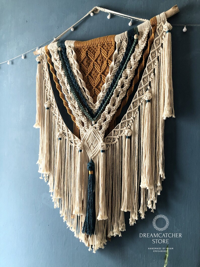 May include: A large, beige macrame wall hanging with a geometric pattern in shades of brown, green, and blue. The hanging is made with thick cotton cord and has a wooden dowel at the top. The hanging is decorated with small white beads and a tassel at the bottom.