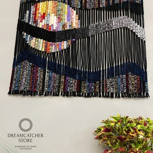 May include: A colorful macrame wall hanging with a black, blue, and white design. The hanging is made of yarn and has a wooden dowel at the top. The text "DREAMCATCHER STORE HANDMADE BY NHAM 0905480089" is at the bottom of the image.