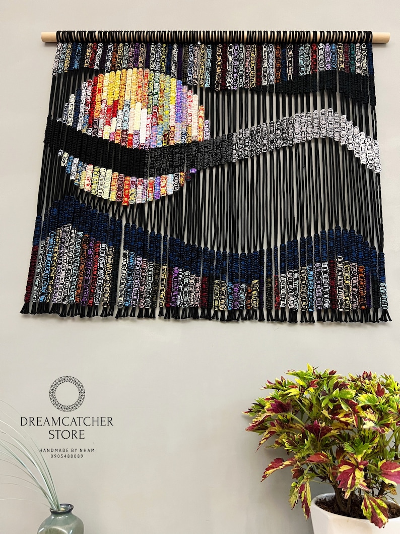 May include: A colorful macrame wall hanging with a black, blue, and white design. The hanging is made of yarn and has a wooden dowel at the top. The text "DREAMCATCHER STORE HANDMADE BY NHAM 0905480089" is at the bottom of the image.