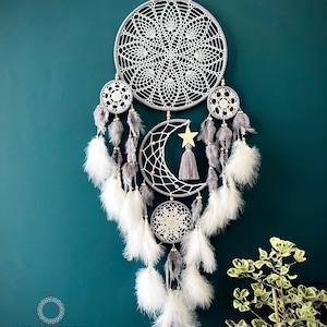 May include: A handcrafted dreamcatcher with a large, circular, crocheted web design. It features smaller circular webs, a crescent moon, and a star, adorned with gray and white feathers. The dreamcatcher hangs against a teal wall.