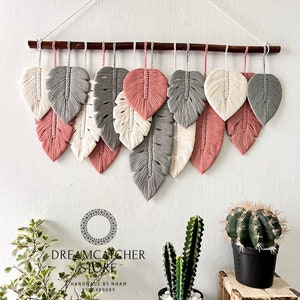 May include: A wall hanging made of macrame feathers in shades of gray, white, and pink. The feathers are arranged in a cascading pattern and are hung from a wooden dowel. The wall hanging is labeled "Dreamcatcher Store" and "Handmade by Nham 0915480089".