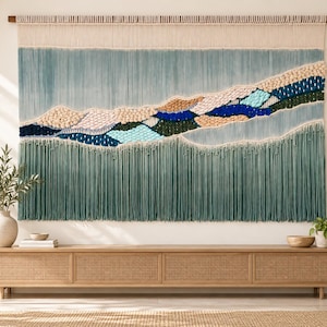 May include: A large macrame wall hanging featuring a landscape design with layers of cream, sage green, dark blue, and light blue fibre. The piece is suspended from a wooden rod and includes a long, dense fringe at the bottom edge.