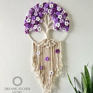 May include: A macrame wall hanging featuring a tree of life design with purple and white crocheted flowers. The hanging has a white background and is made with natural fibers.