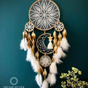 May include: A large, white crocheted dreamcatcher with brown feathers and beads hanging from it. The dreamcatcher is decorated with a crescent moon and a star. The dreamcatcher is hanging on a teal wall.