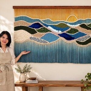 Peut inclure: Une grande tenture murale en macramé rectangulaire présentant un paysage aux textures superposées dans des tons de jaune, bleu, vert et beige. L'œuvre est fixée sur une tige en bois foncé et comporte une longue frange fluide en bas.
