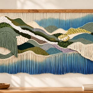 May include: A large macrame wall hanging featuring a landscape design with layered textures in shades of sage green, forest green, cream, and blue. The piece is mounted on a wooden rod and includes long, flowing fringe at the bottom edge.