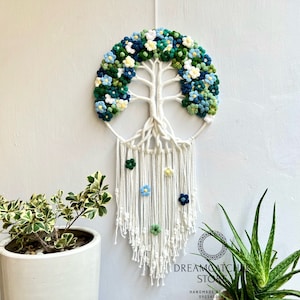 May include: A white macrame dreamcatcher with a crocheted tree of life design. The tree is decorated with green and blue flowers. The dreamcatcher has white tassels and is hanging on a white wall.