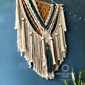 May include: A large macrame wall hanging with a geometric design in shades of beige, mustard yellow, teal, and green. The hanging has a fringe of white tassels and is mounted on a wooden dowel.