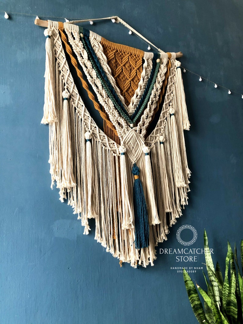 May include: A large macrame wall hanging with a geometric design in shades of beige, mustard yellow, teal, and green. The hanging has a fringe of white tassels and is mounted on a wooden dowel.