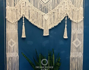 May include: A white macrame wall hanging with a geometric design. The hanging is made of cotton cord and has a fringe border. The hanging is displayed against a blue wall with a green plant in front of it. The text "DREAMCATCHER STORE HANDMADE BY NHAM 0905480889" is printed below the hanging.