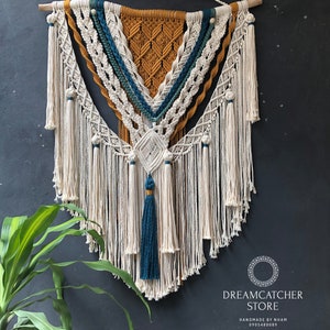 May include: A large macrame wall hanging with a geometric design in shades of white, mustard yellow, and teal. The hanging features a fringe border and a tassel at the bottom. The text "DREAMCATCHER STORE HANDMADE BY NHAM 0905480089" is visible at the bottom of the image.