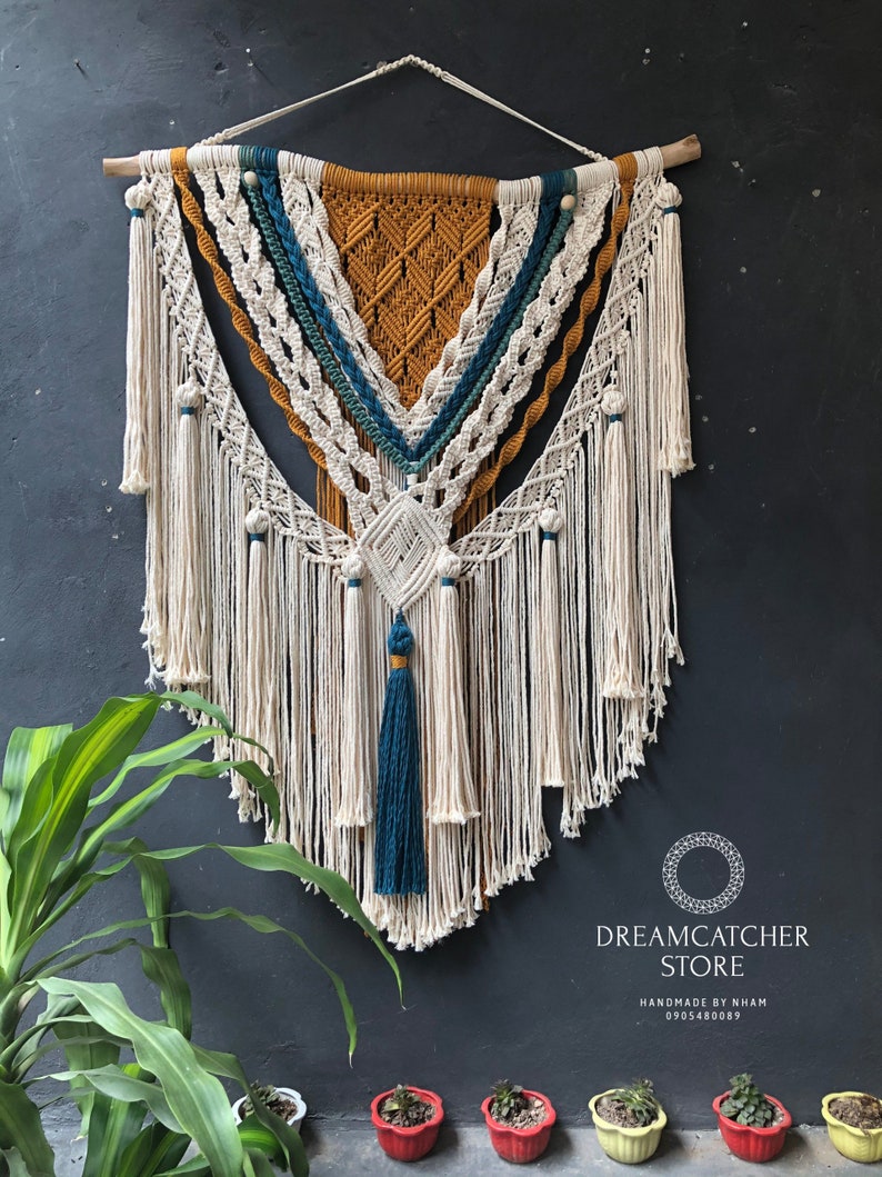 May include: A large macrame wall hanging with a geometric design in shades of white, mustard yellow, and teal. The hanging features a fringe border and a tassel at the bottom. The text "DREAMCATCHER STORE HANDMADE BY NHAM 0905480089" is visible at the bottom of the image.