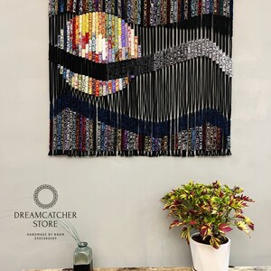 May include: A black string wall hanging with a colorful, abstract design. The design features a circle with a wave-like pattern. The wall hanging is made of yarn and is hung on a wooden dowel.