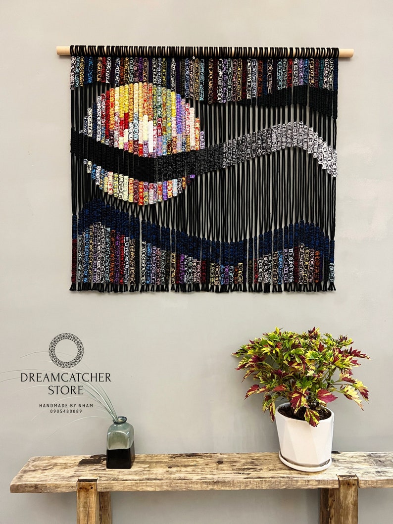 May include: A black string wall hanging with a colorful, abstract design. The design features a circle with a wave-like pattern. The wall hanging is made of yarn and is hung on a wooden dowel.