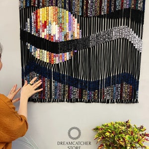 May include: A colorful macrame wall hanging with a black and white design. The hanging is made with a variety of colored cords and beads. The hanging is titled "Dreamcatcher Store" and includes the text "Handmade by Nham 0905480089".