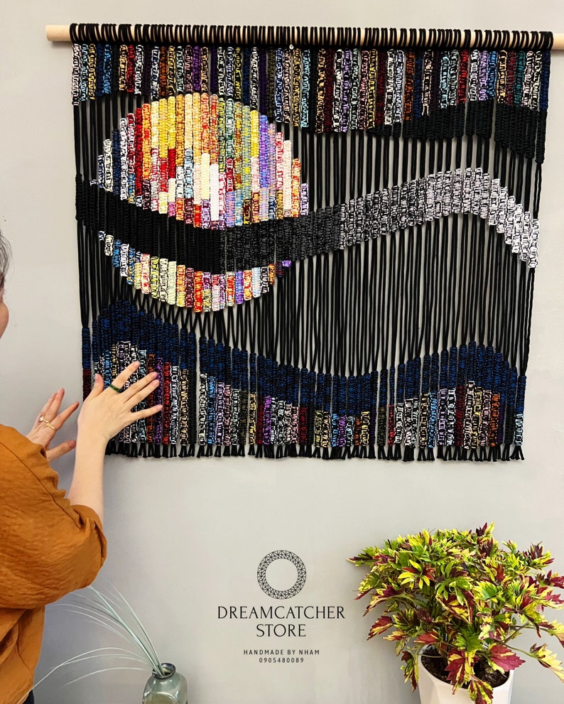 May include: A colorful macrame wall hanging with a black and white design. The hanging is made with a variety of colored cords and beads. The hanging is titled "Dreamcatcher Store" and includes the text "Handmade by Nham 0905480089".