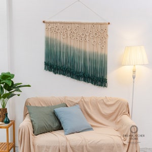 May include: A green and white ombre macrame wall hanging with a geometric pattern of circles. The hanging is made of cotton cord and is approximately 24 inches wide and 36 inches long. The hanging is displayed on a white wall above a beige couch with green and blue throw pillows.