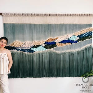 Puede incluir: Tapiz de pared hecho a mano con un diseño de paisaje montañoso. El tapiz utiliza una variedad de texturas y colores, incluyendo beige, azul y verde, para crear un efecto tridimensional. Está montado sobre una barra de madera.