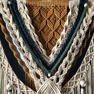 May include: A macrame wall hanging with a geometric design in beige, mustard yellow, teal, and dark blue. The hanging features a variety of knots and tassels.