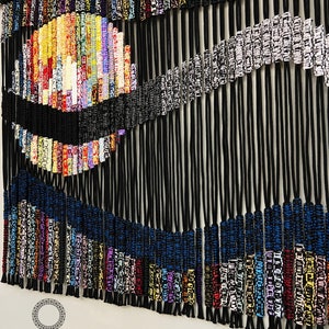 May include: A colorful macrame wall hanging with black, blue, white, and multi-colored cords. The wall hanging features a circular design with a wave-like pattern. The text "DREAMCATCHER STORE HANDMADE BY NHAM 0905480089" is at the bottom of the image.