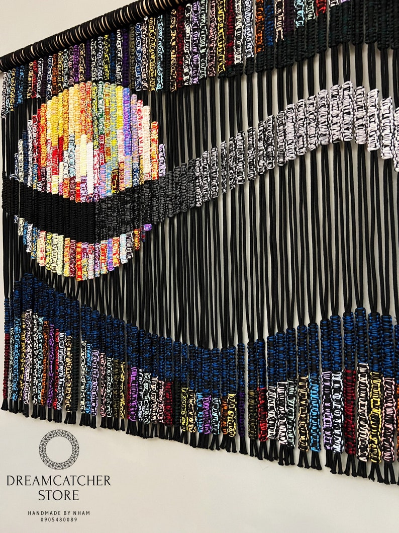 May include: A colorful macrame wall hanging with black, blue, white, and multi-colored cords. The wall hanging features a circular design with a wave-like pattern. The text "DREAMCATCHER STORE HANDMADE BY NHAM 0905480089" is at the bottom of the image.